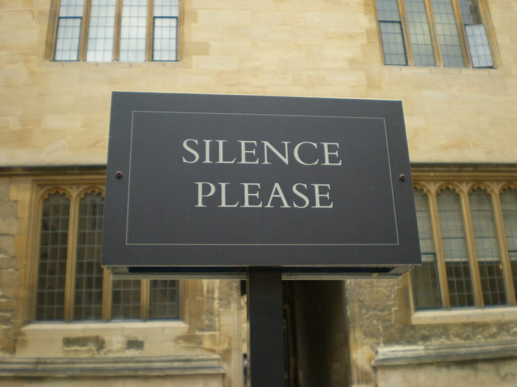 A dark rectangular sign reading “Silence please” in pale capital letters outside a stone building.